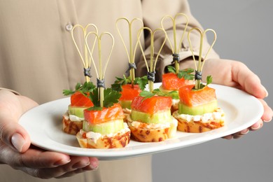 Photo of Woman holding tasty canapes with salmon, cucumber, bread and cream cheese on grey background, closeup