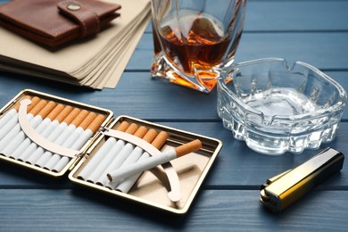 Photo of Open case with tobacco filter cigarettes, lighter, ashtray and alcohol drink on blue wooden table, closeup