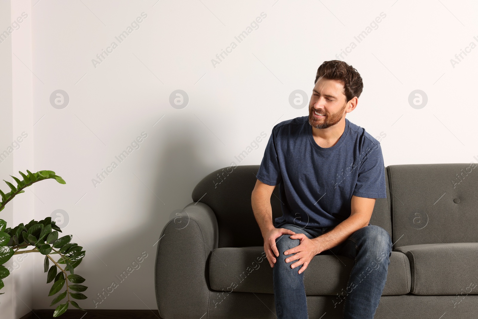 Photo of Man suffering from leg pain and touching knee on sofa indoors, space for text