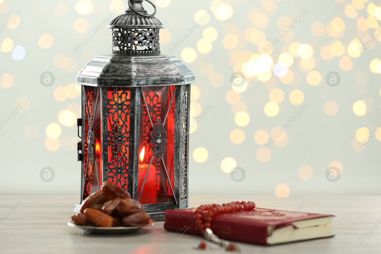 Photo of Arabic lantern, Quran, misbaha and dates on table against blurred lights