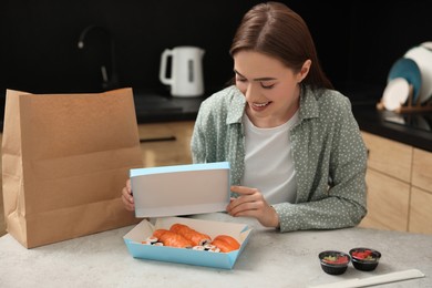 Photo of Beautiful young woman unpacking her order from sushi restaurant at table in kitchen