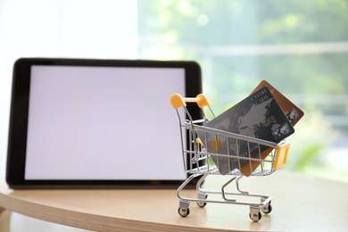 Internet shopping. Small cart with credit cards near modern tablet on table indoors