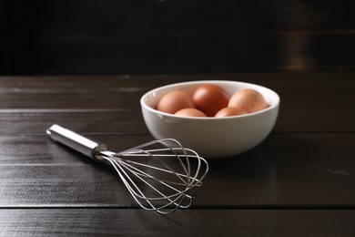 Metal whisk and raw eggs in bowl on dark wooden table, closeup