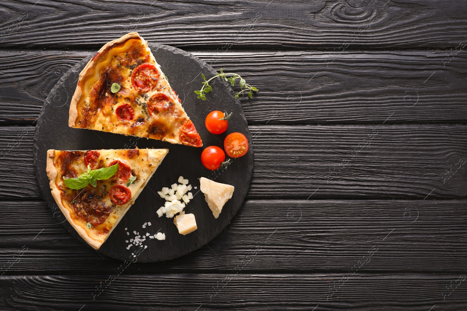 Photo of Pieces of delicious homemade quiche with prosciutto and ingredients on black wooden table, flat lay. Space for text