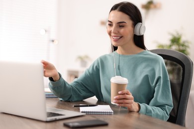 Photo of Young woman in headphones using video chat during webinar at table in room