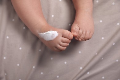 Photo of Cute little baby with moisturizing cream on her foot in bed, closeup