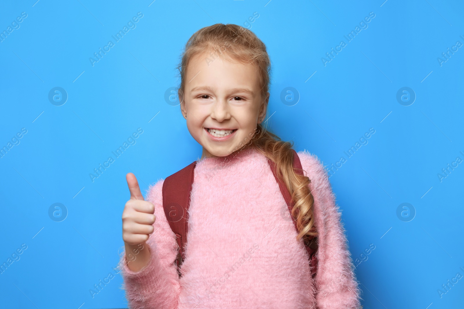 Photo of Happy little girl with backpack showing thumbs up on light blue background