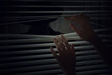 Woman opening window blinds and looking at stranger in darkness, closeup. Paranoia concept