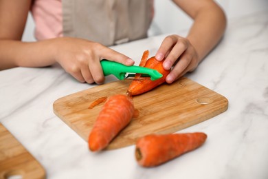 Photo of Little girl peeling carrot at table in kitchen, closeup. Preparing vegetable