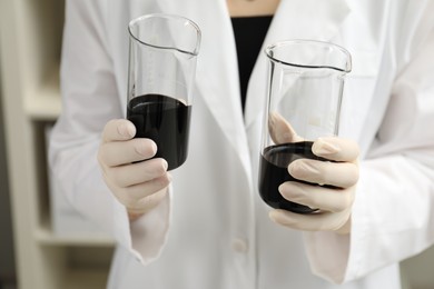 Photo of Laboratory worker holding beakers with black crude oil indoors, closeup