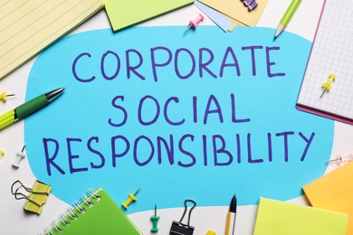 Photo of Paper text balloon with phrase Corporate Social Responsibility among office supplies on white table, flat lay