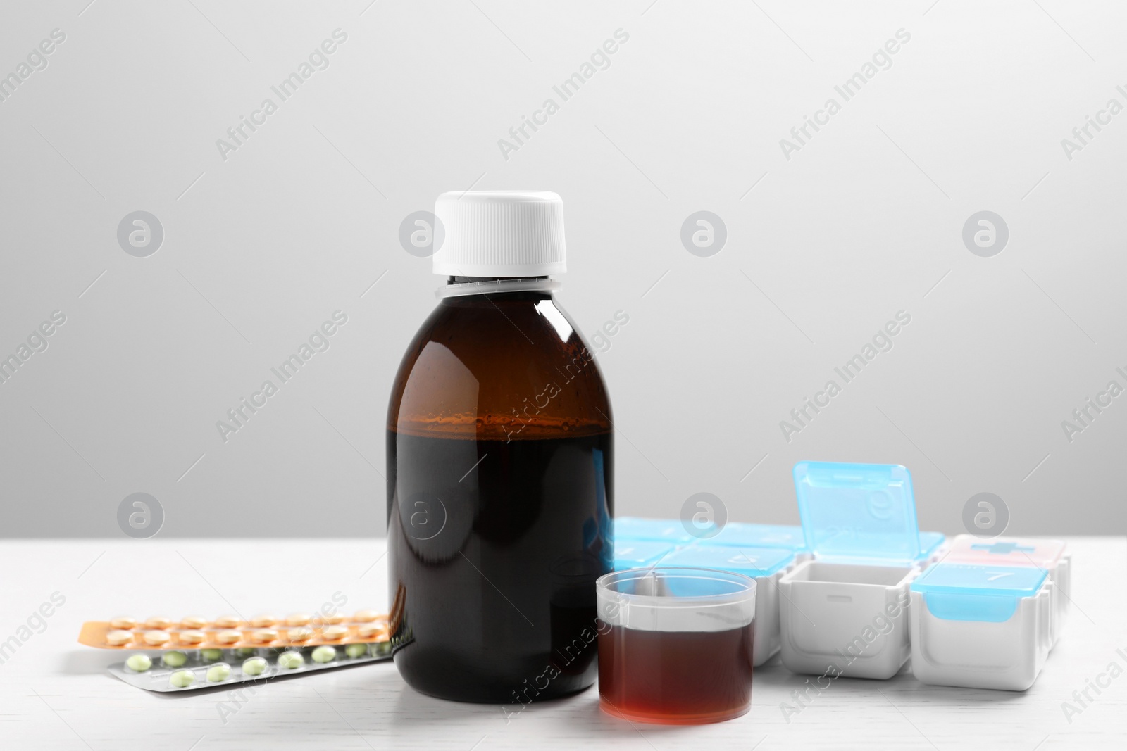 Photo of Bottle of syrup, measuring cup, weekly pill organizer and pills on white table against light grey background. Cold medicine