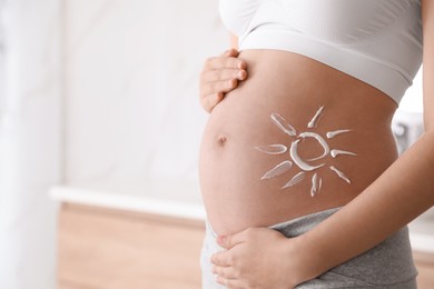 Photo of Young pregnant woman with sun protection cream on belly indoors, closeup. Space for text