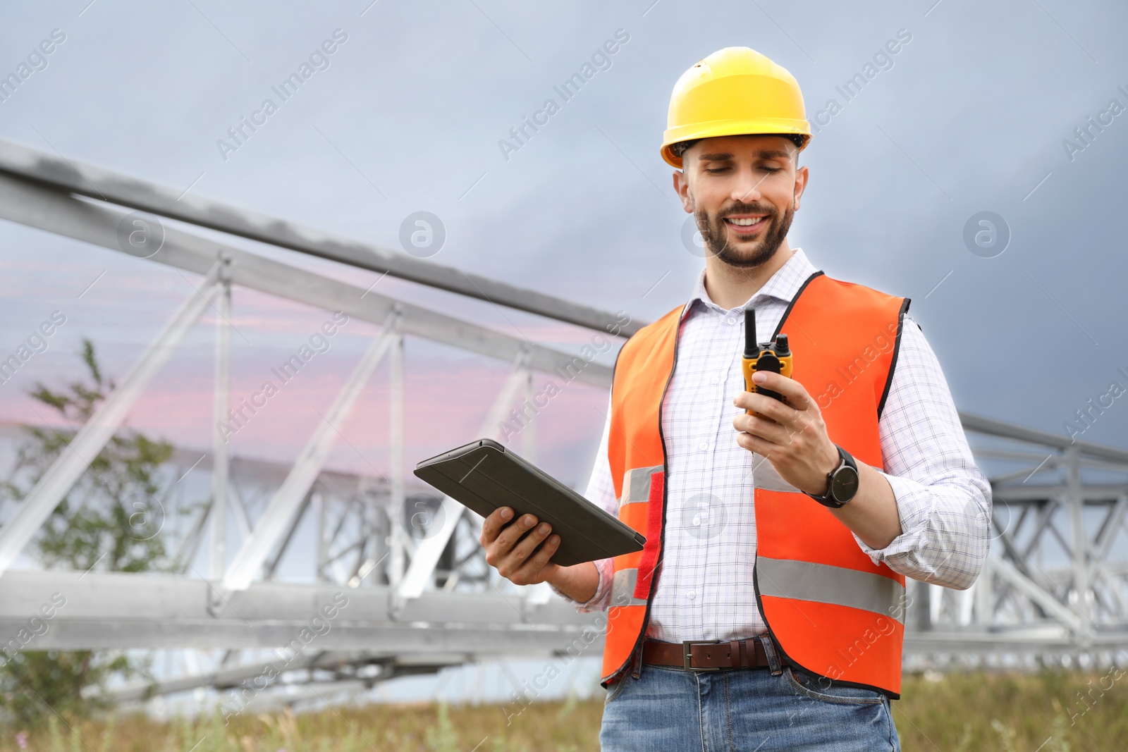 Photo of Professional engineer with tablet and walkie talkie near high voltage tower construction outdoors. Installation of electrical substation