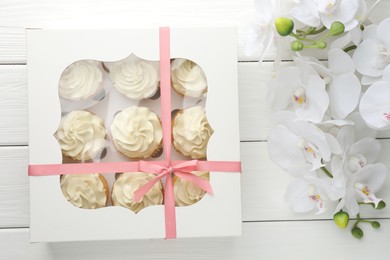 Photo of Tasty cupcakes with vanilla cream in box and orchid flowers on white wooden table, top view