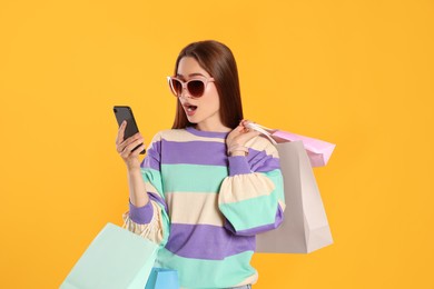 Surprised young woman with shopping bags looking at smartphone on yellow background. Big sale