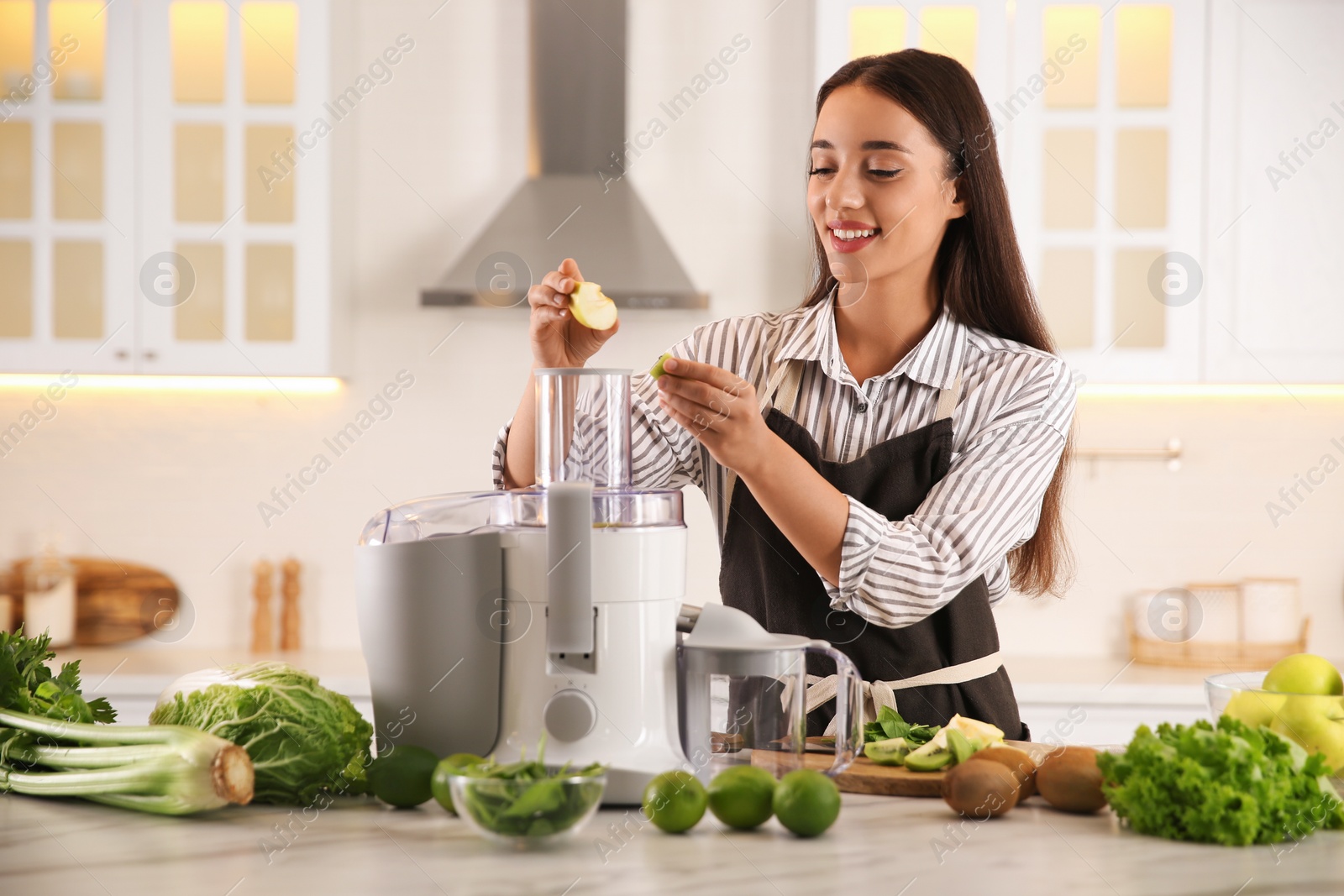 Photo of Young woman putting fresh kiwi and apple into juicer at table in kitchen