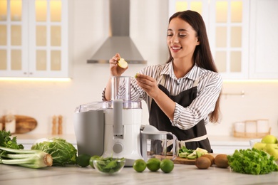 Photo of Young woman putting fresh kiwi and apple into juicer at table in kitchen