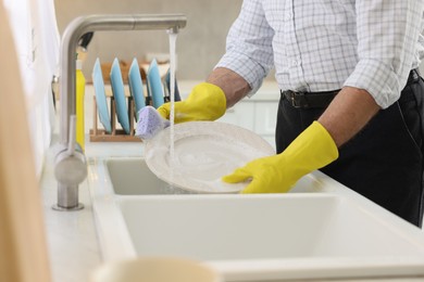 Man in protective gloves washing plate above sink in kitchen, closeup