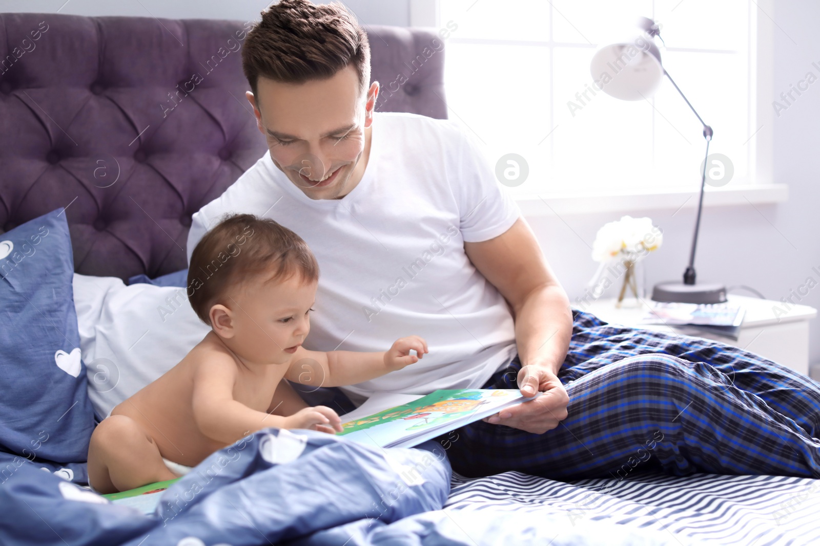 Photo of Young father reading book with his cute little son on bed