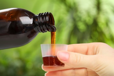Photo of Woman pouring syrup from bottle into measuring cup on blurred background, closeup. Cold medicine