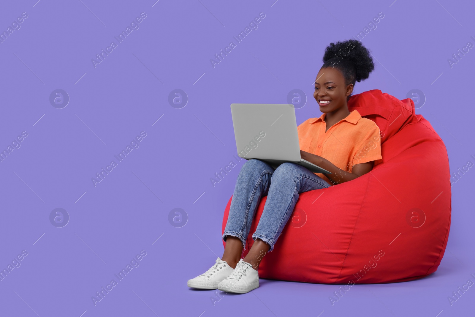 Photo of Beautiful young woman with laptop sitting on beanbag chair against purple background. Space for text