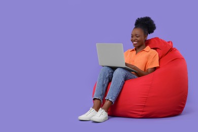 Beautiful young woman with laptop sitting on beanbag chair against purple background. Space for text