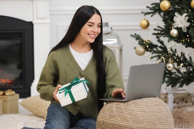 Photo of Celebrating Christmas online with exchanged by mail presents. Smiling woman with gift box during video call on laptop at home