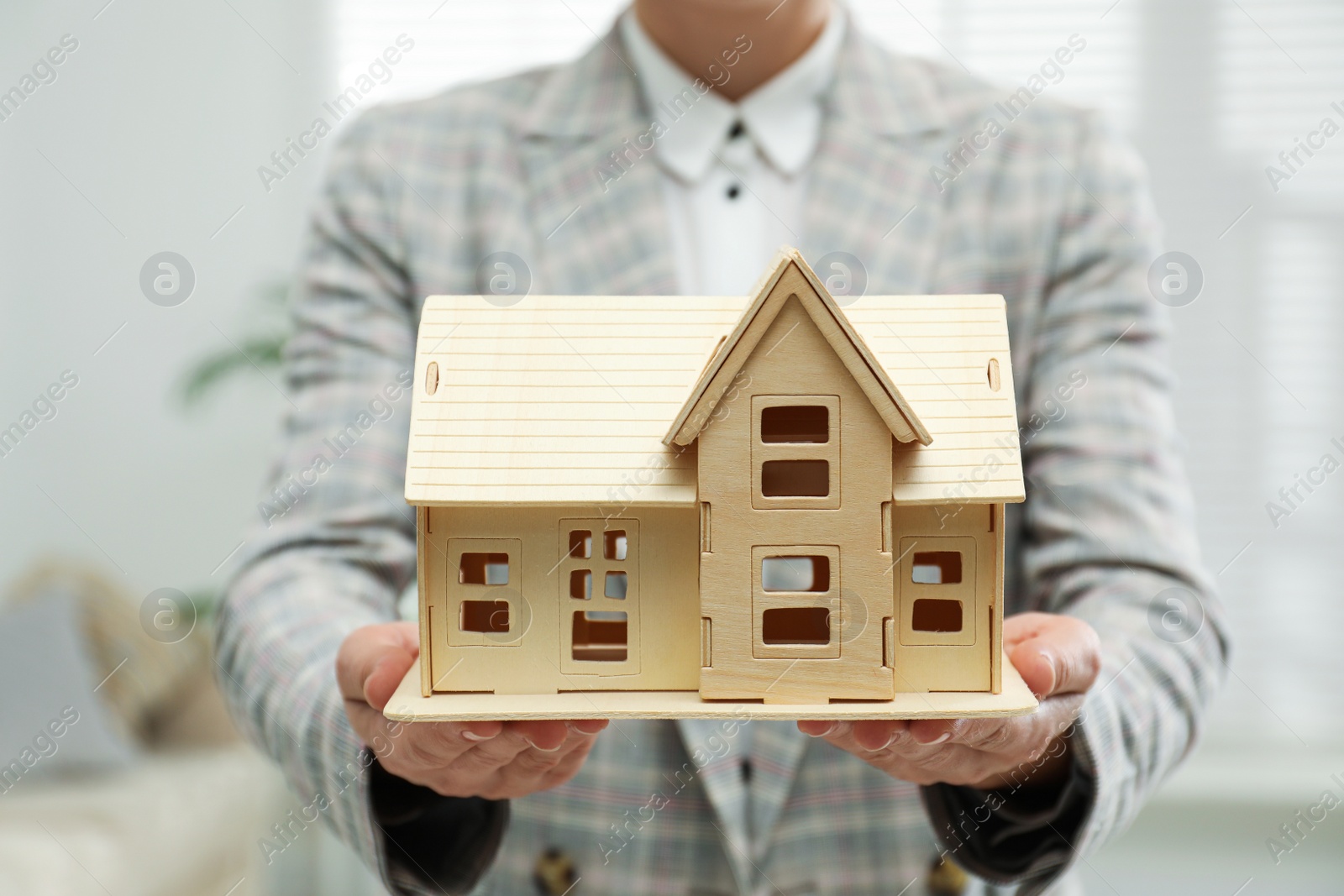 Photo of Real estate agent holding wooden house model indoors, closeup