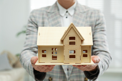 Photo of Real estate agent holding wooden house model indoors, closeup