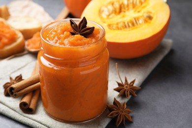 Photo of Jar of pumpkin jam, star anise, cinnamon and fresh pumpkin on grey table, closeup