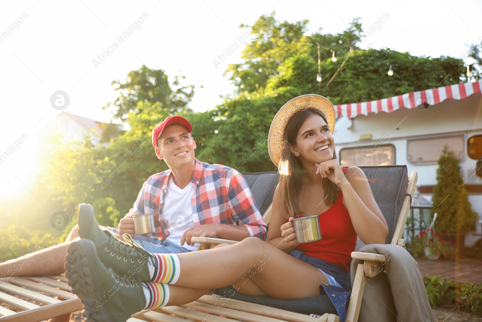 Photo of Happy couple with cups resting on deck chairs near motorhome. Camping season