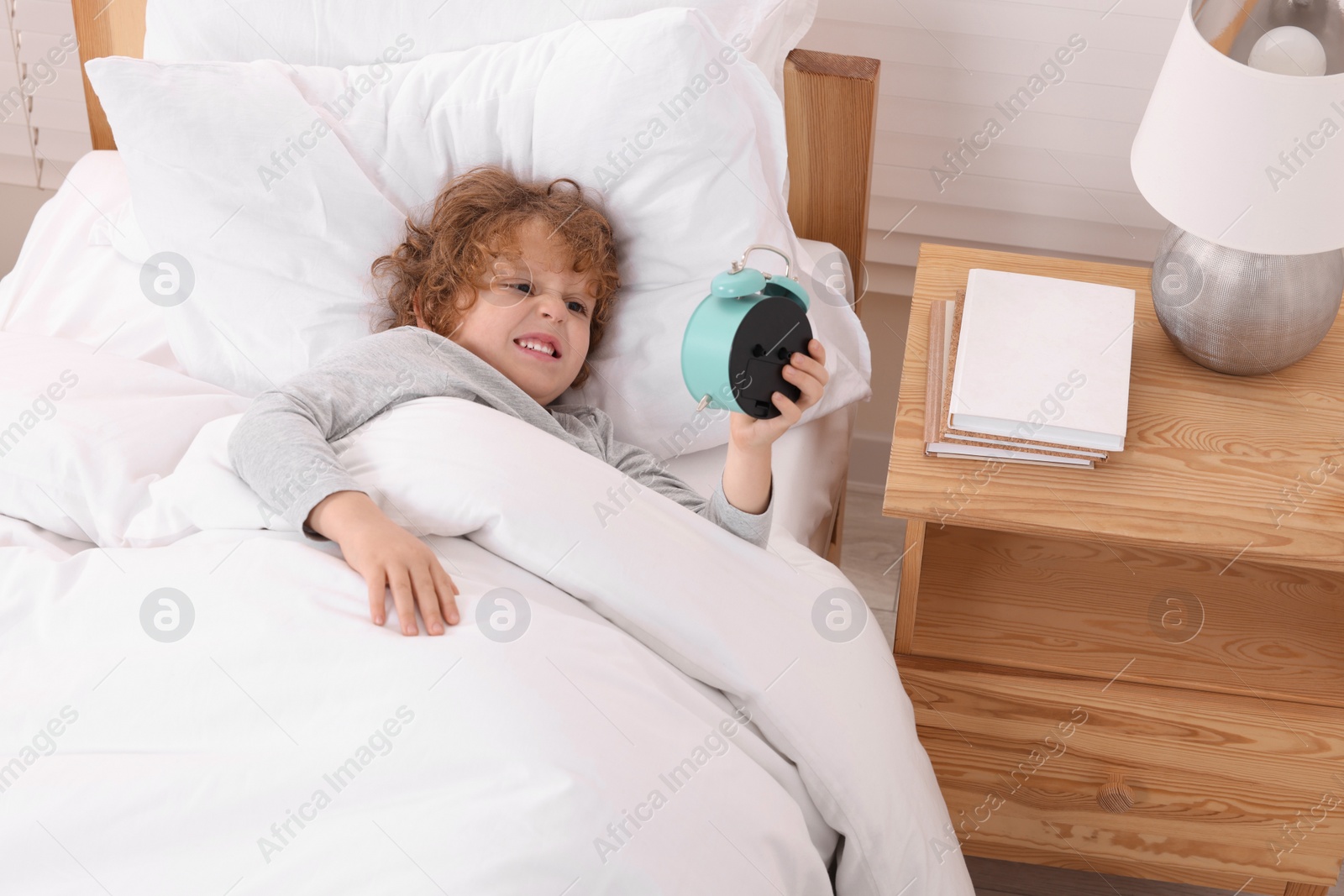 Photo of Emotional little boy looking at alarm clock in bedroom