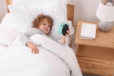 Photo of Emotional little boy looking at alarm clock in bedroom