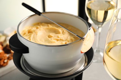 Photo of Fork with piece of apple, melted cheese in fondue pot and wine on grey table, closeup