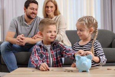 Photo of Family budget. Children putting coins into piggy bank and their parents at table indoors, selective focus