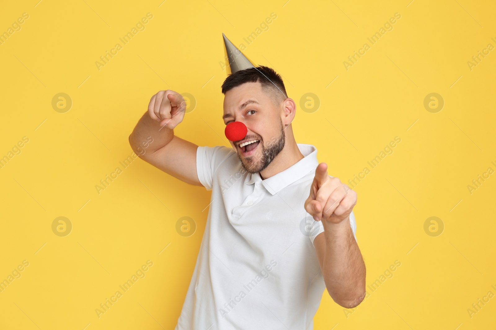 Photo of Funny man with clown nose and party hat on yellow background. April fool's day