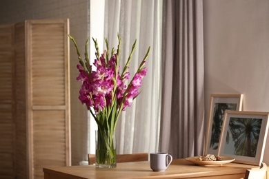 Photo of Vase with beautiful pink gladiolus flowers, pictures and cup on wooden table in room, space for text