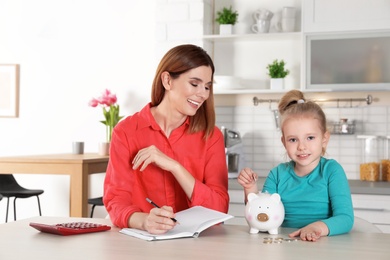 Mother and daughter putting coin into piggy bank at table indoors. Saving money