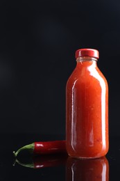 Photo of Spicy chili sauce in bottle and pepper against dark background, space for text