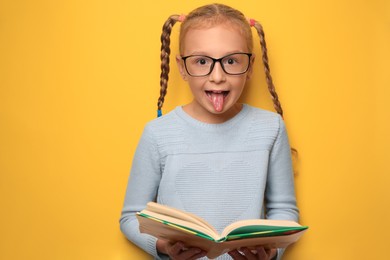 Photo of Cute little girl with glasses and textbook on yellow background