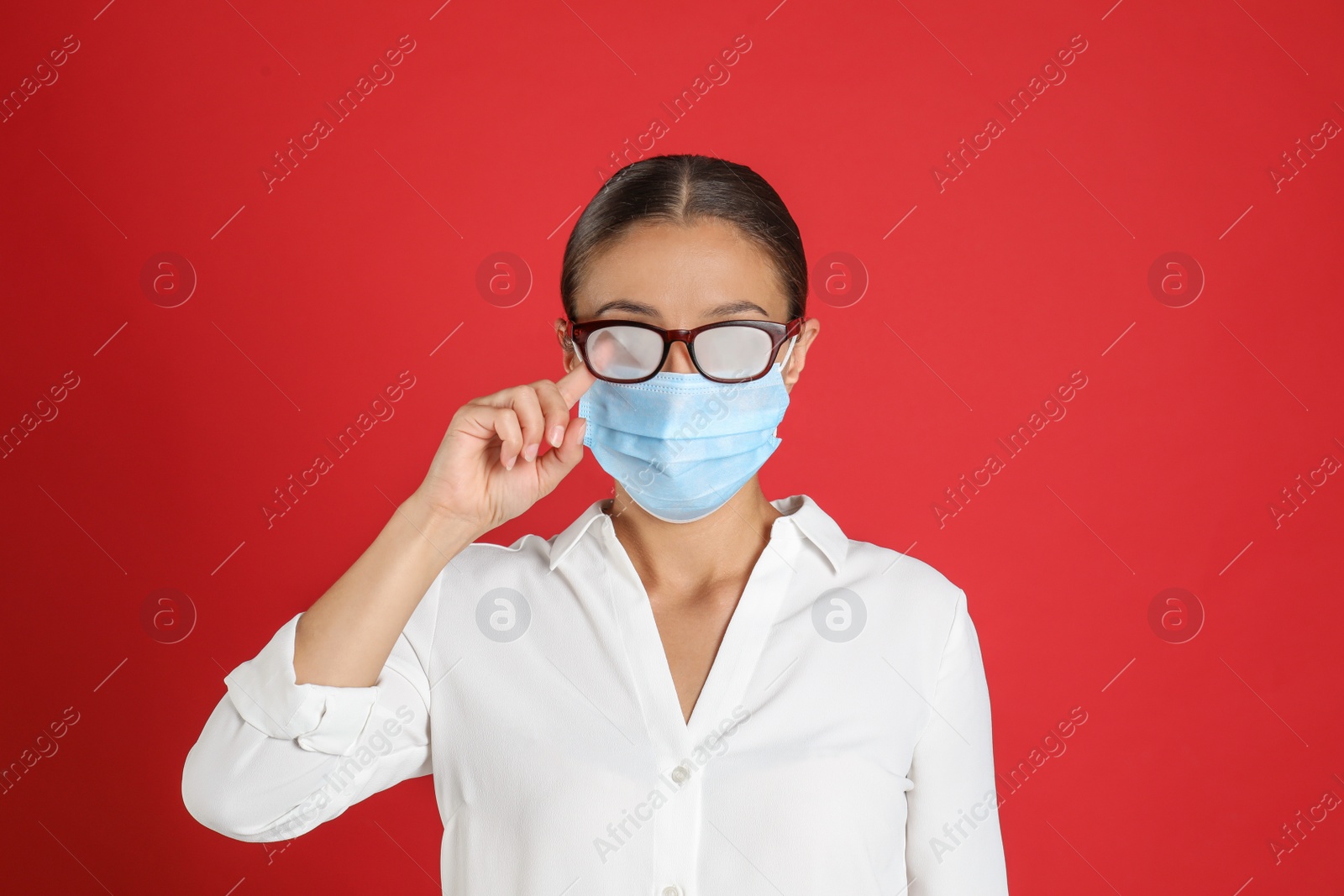 Photo of Woman wiping foggy glasses caused by wearing medical mask on red background