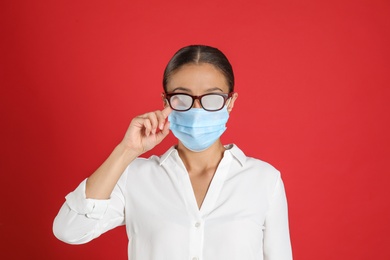 Photo of Woman wiping foggy glasses caused by wearing medical mask on red background