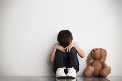 Photo of Sad little boy with toy sitting near white wall, space for text