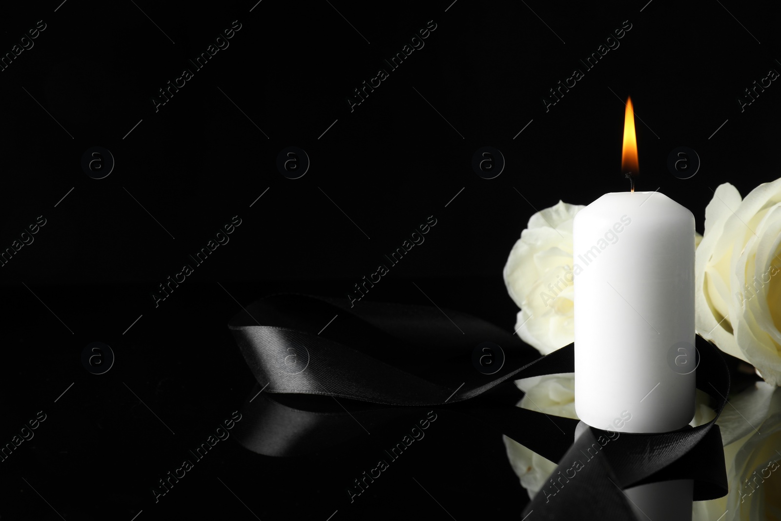 Photo of Burning candle, white roses and ribbon on black mirror surface in darkness, closeup with space for text. Funeral symbols