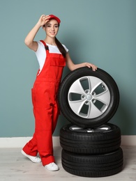 Female mechanic in uniform with car tires on color wall background