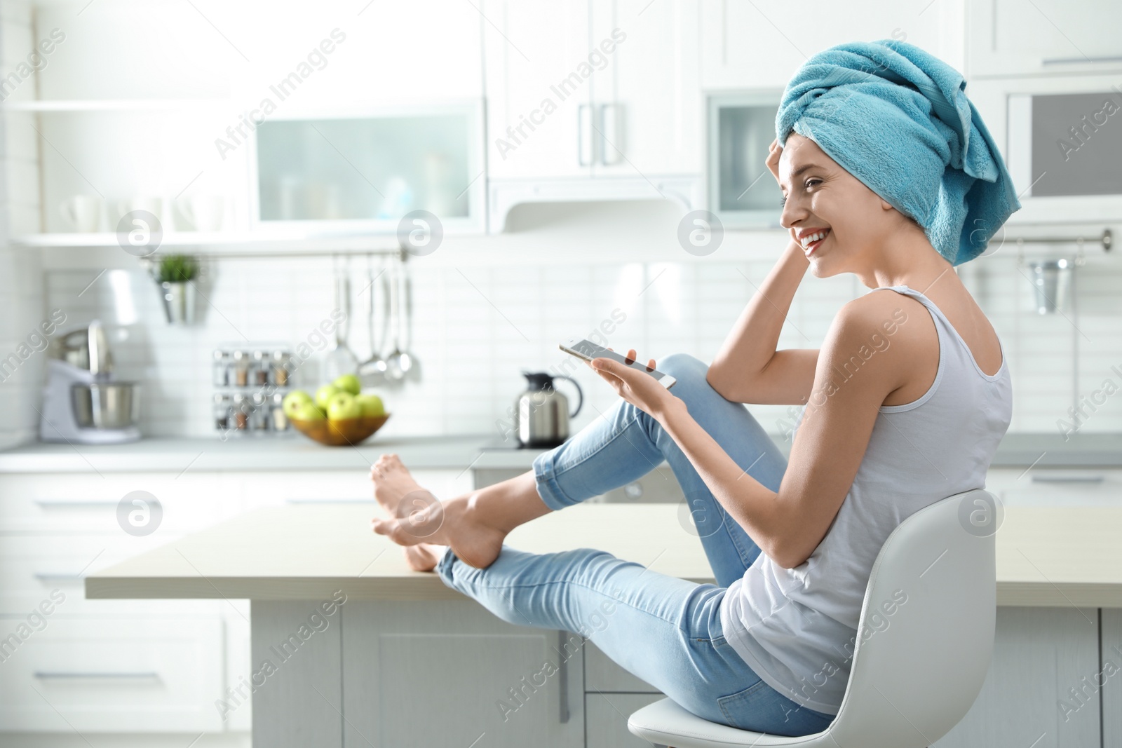 Photo of Pretty young woman with towel on head using smartphone in kitchen