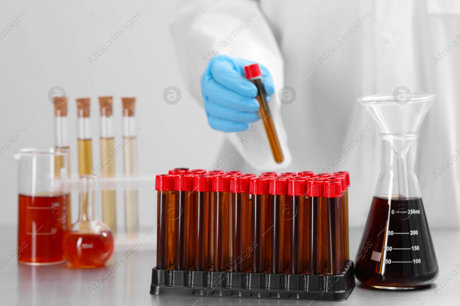 Photo of Scientist holding test tube with brown liquid near different laboratory glassware on table, selective focus
