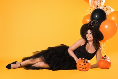 Photo of Beautiful woman in witch costume with balloons and pumpkins on yellow background, space for text. Halloween party