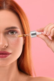 Beautiful young woman applying cosmetic serum onto her face on pink background, closeup